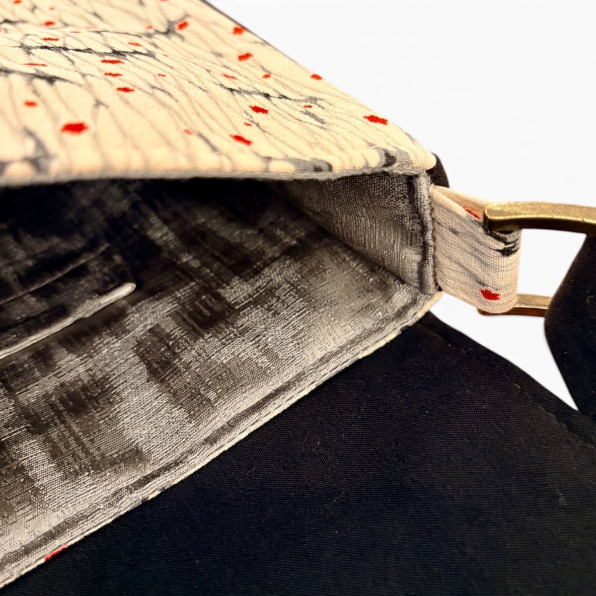 Close-up of a fabric bag corner showing a cream and black marbled pattern with small red speckles, a black velvet-like interior lining, and a gold-tone metal ring attachment.
