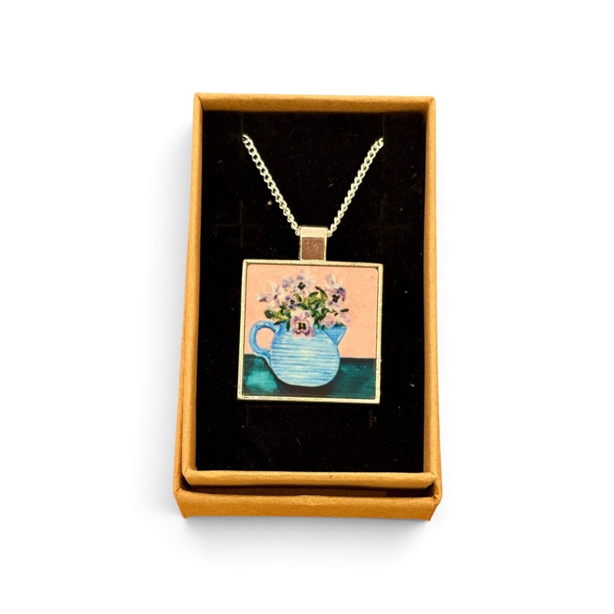 A square pendant necklace displayed in an open tan gift box with a black velvet insert. The pendant features a colorful illustration of a blue teapot filled with flowers against a peach and teal background, suspended from a silver-toned chain.