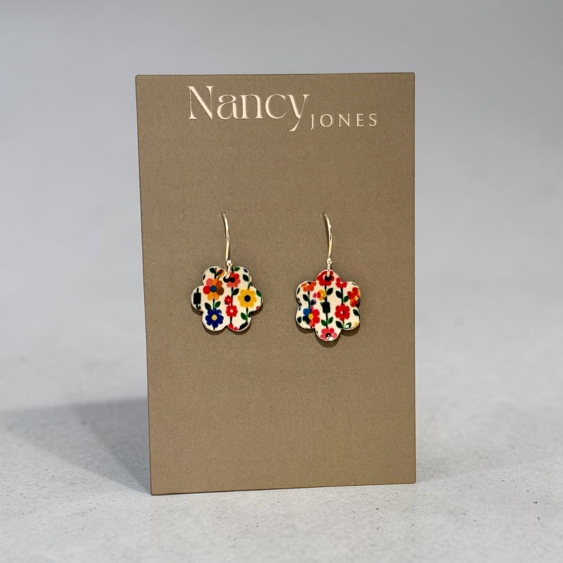 A pair of Nancy Jones floral enamel flower-shaped drop earrings displayed on a brown cardboard brand card against a neutral gray background.