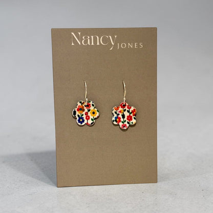 A pair of Nancy Jones floral enamel flower-shaped drop earrings displayed on a brown cardboard brand card against a neutral gray background.