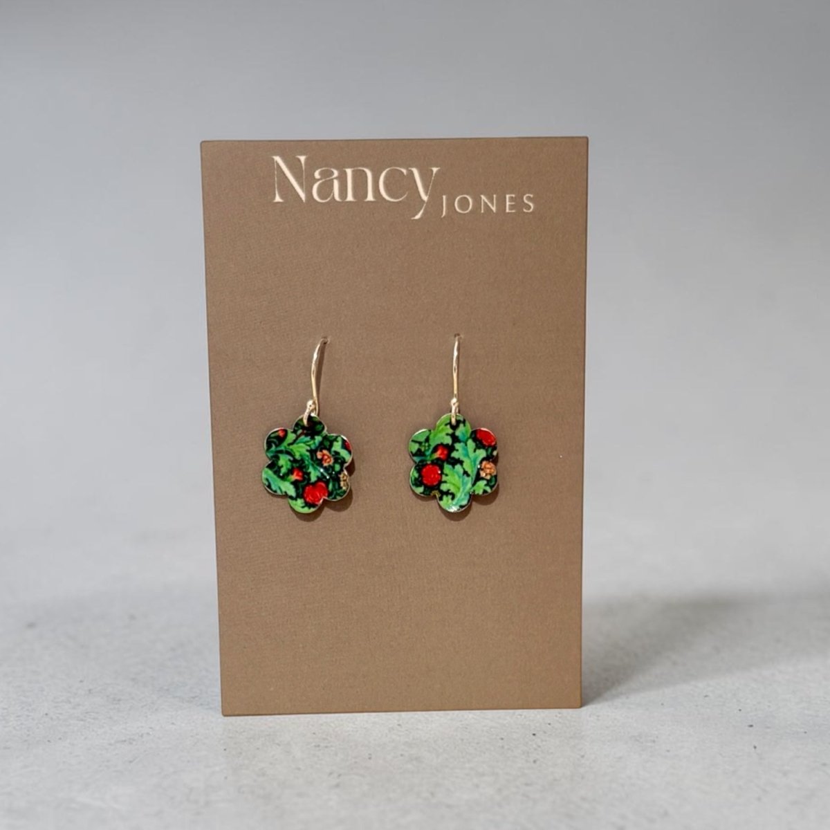 A pair of Nancy Jones dangle earrings with green floral clusters and red berries, displayed on a brown cardboard backing card against a neutral gray background.
