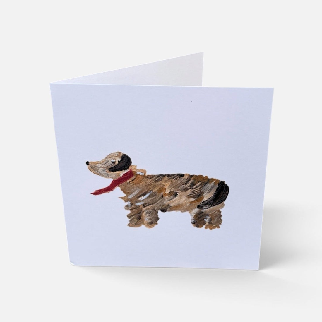 A folded greeting card standing upright on a light gray background. The front of the card features a painted dachshund in brown, black, and tan with a red scarf on a pale lavender card background.
