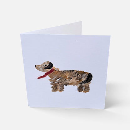 A folded greeting card standing upright on a light gray background. The front of the card features a painted dachshund in brown, black, and tan with a red scarf on a pale lavender card background.