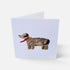 A folded greeting card standing upright on a light gray background. The front of the card features a painted dachshund in brown, black, and tan with a red scarf on a pale lavender card background.