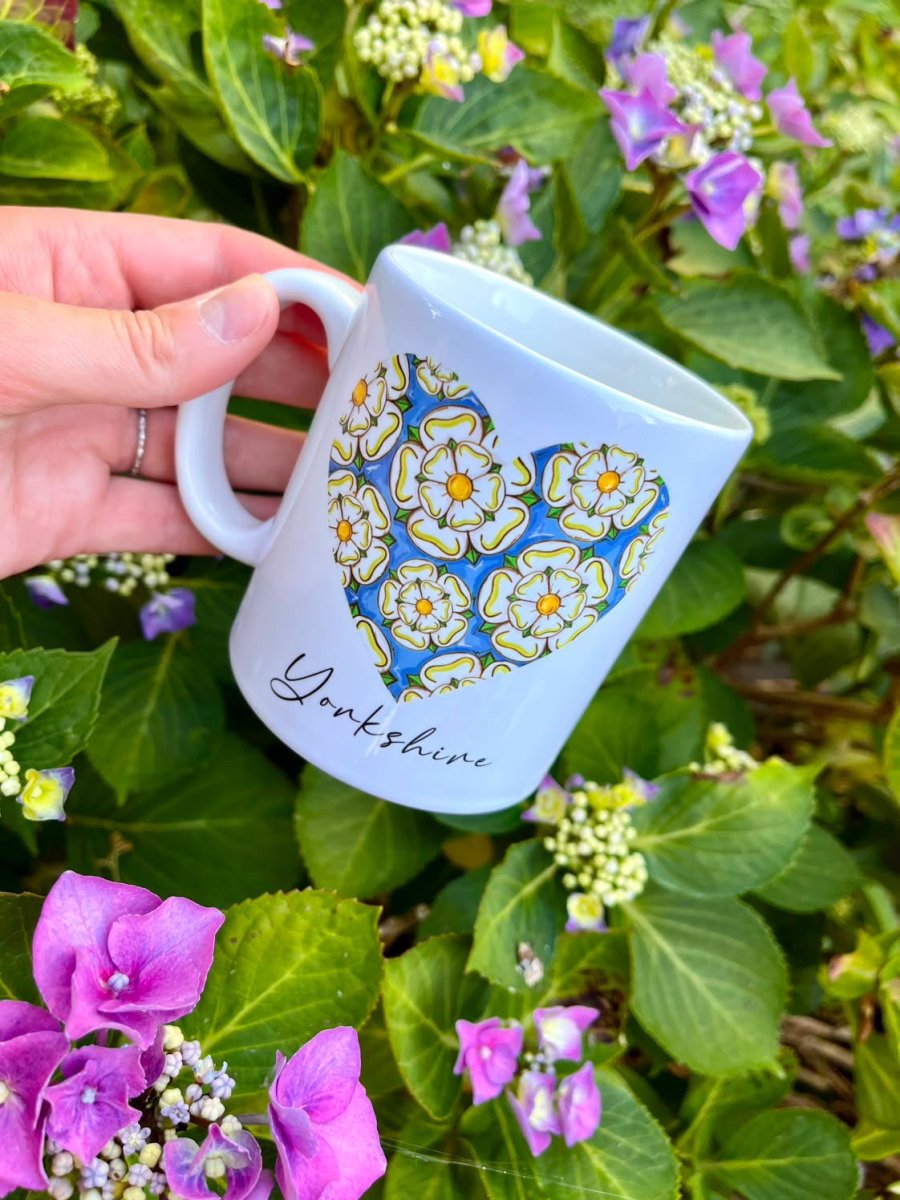 A hand holds a white ceramic mug decorated with a blue and yellow floral heart design and the word &